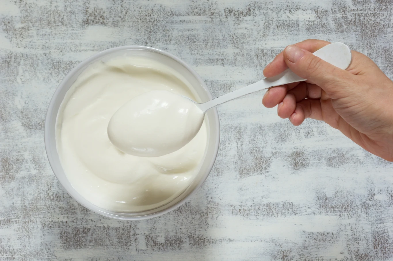 Jogurt z dodatkiem czarnej rzepy będzie świetnym pomysłem na domową maseczkę Plastikowa łyżka nabiera gęsty, biały jogurt z przezroczystej miski ustawionej na jasnym tle.