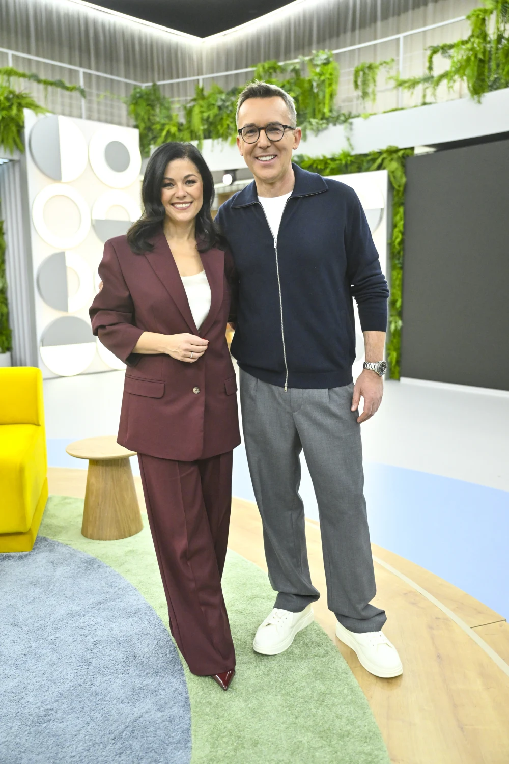 Katarzyna Cichopek w garniturze idealnym na święta Dwoje uśmiechniętych prowadzących pozuje razem w nowoczesnym, jasnym studiu telewizyjnym z zielonymi elementami dekoracyjnymi i kolorowymi meblami.