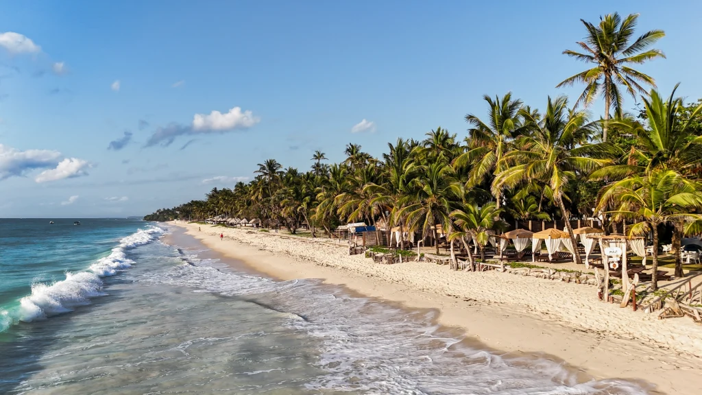 Szeroka, piaszczysta plaża otoczona wysokimi palmami kokosowymi. Delikatne fale uderzają o brzeg, a w tle po prawej stronie widać zacienione strefy wypoczynku i altany. Całość rozświetlona jest łagodnym, naturalnym światłem, a niebo ozdobione jest kilk...