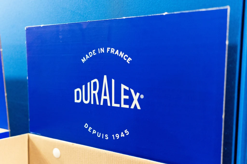 Marka Duralex miała ostatnio spore kłopoty Niebieskie pudełko z dużym białym napisem Duralex, informacją Made in France oraz datą powstania marki 1945, ukazane na tle ściany w podobnym kolorze.