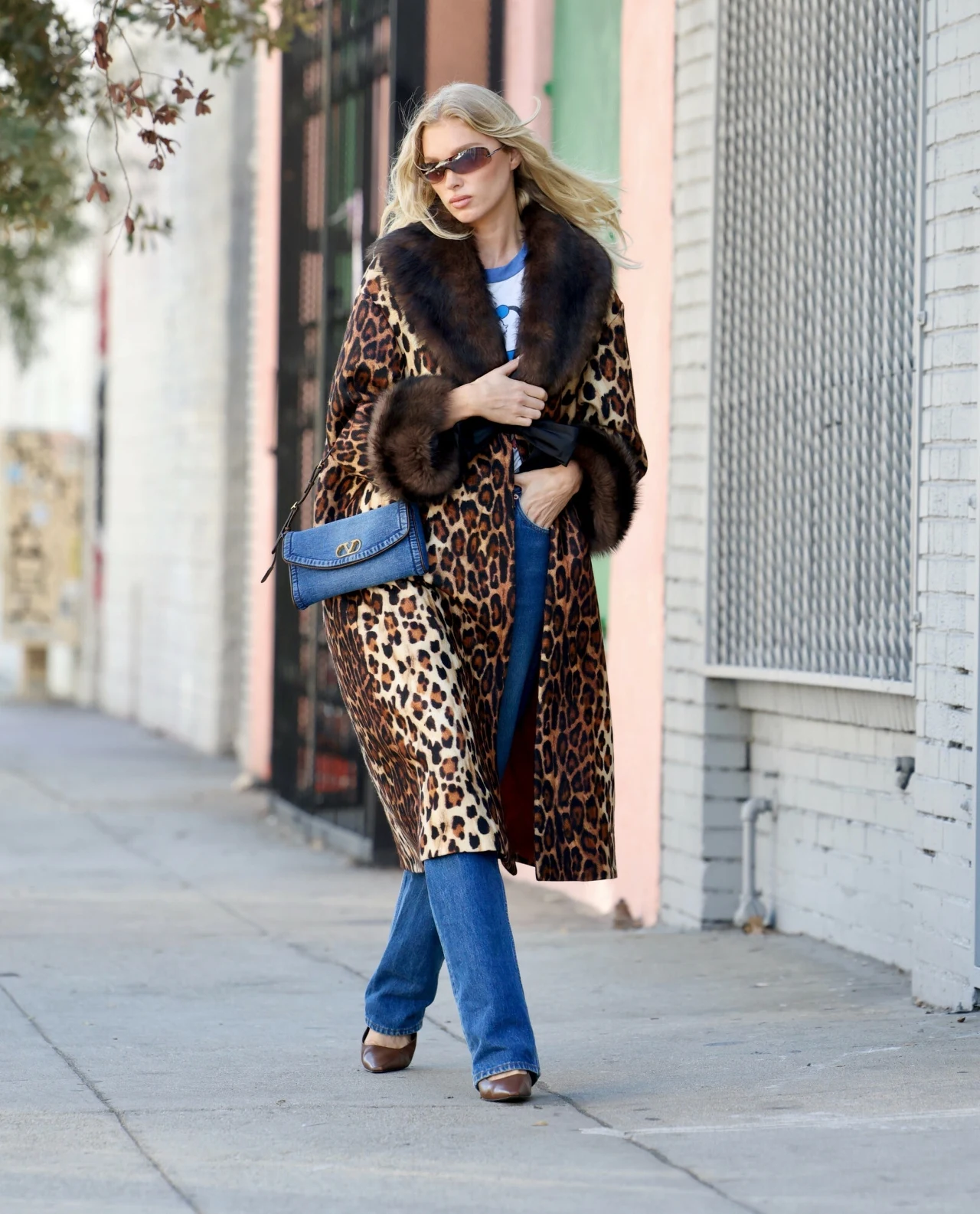 Elsa Hosk w panterkowym płaszczu Kobieta w długim płaszczu w lamparcie cętki z brązowym futrzanym kołnierzem i mankietami, niebieska torebka przewieszona przez ramię, niebieskie dżinsy i brązowe buty na obcasie, jasne włosy, okulary przeciwsłoneczne, idzie chodnikiem przy miejskiej ul...