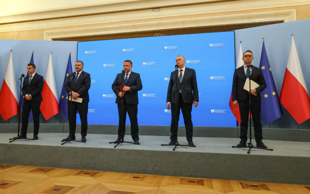 Pięciu mężczyzn w garniturach stoi na podium przed mikrofonami, na tle niebieskiej ścianki z logotypami rządowymi oraz flagami Polski i Unii Europejskiej po bokach.