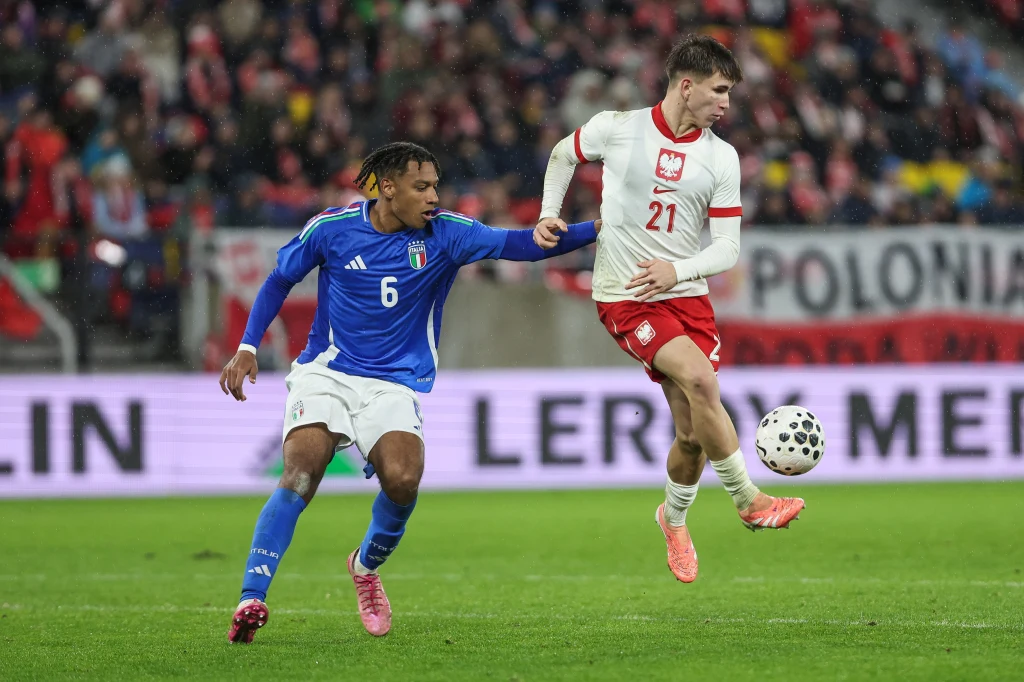 Dwóch piłkarzy rywalizuje o piłkę podczas meczu, zawodnik w niebieskiej koszulce reprezentuje Włochy, a w białej i czerwonej – Polskę, w tle kibice na trybunach.