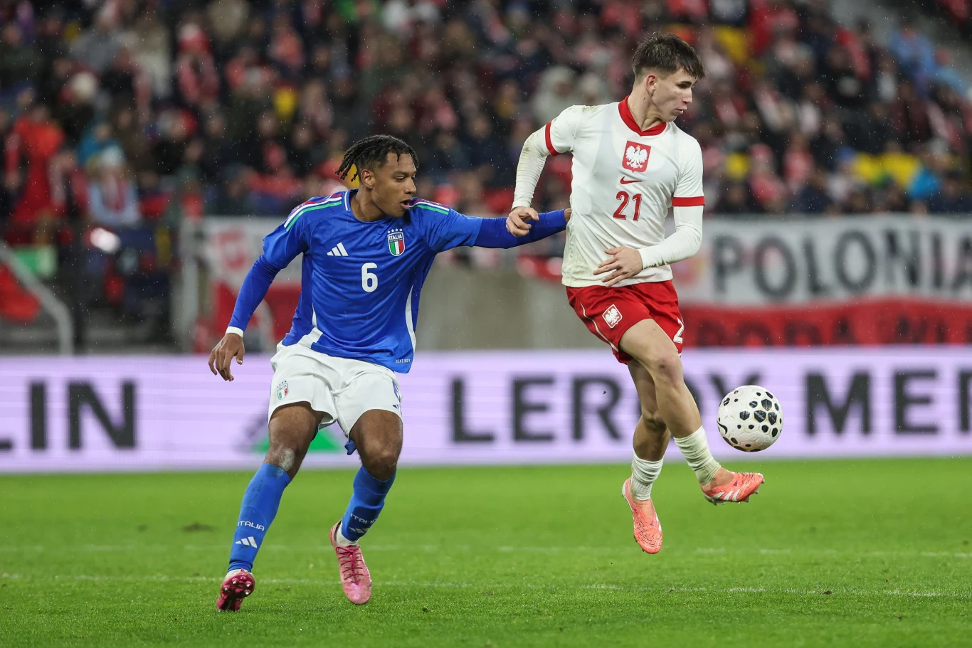 Marcel Reguła Dwóch piłkarzy rywalizuje o piłkę podczas meczu, zawodnik w niebieskiej koszulce reprezentuje Włochy, a w białej i czerwonej – Polskę, w tle kibice na trybunach.