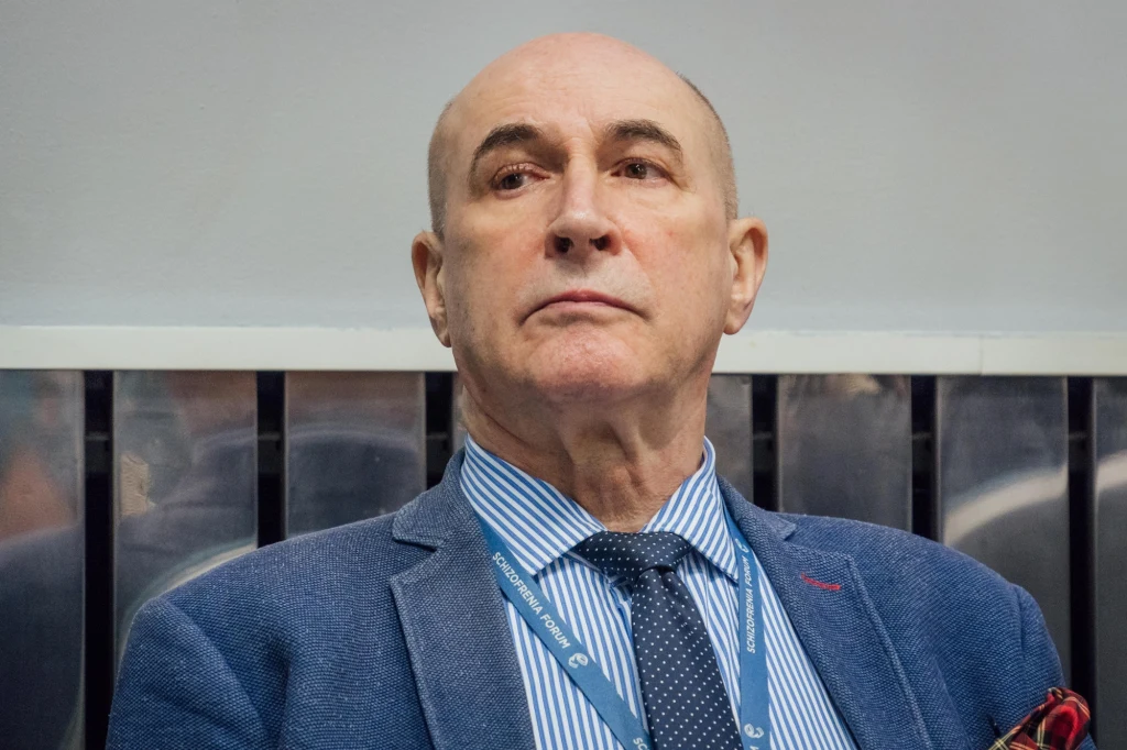 Prof. Janusz Heitzman Starszy mężczyzna w niebieskiej marynarce, jasnoniebieskiej koszuli w paski i granatowym krawacie siedzi na tle metalowej kratownicy; na szyi zawieszony identyfikator.