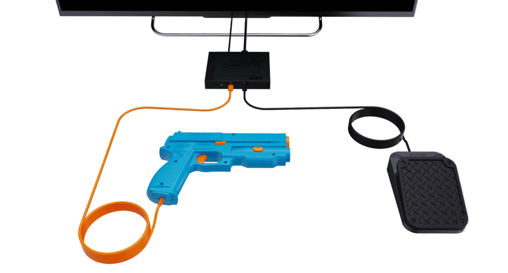 Plastikowy pistolet w kolorze niebieskim z pomarańczowym przewodem, podłączony do czarnego urządzenia oraz pedału, całość leży na jasnym tle i jest elementem elektronicznej gry lub symulatora.