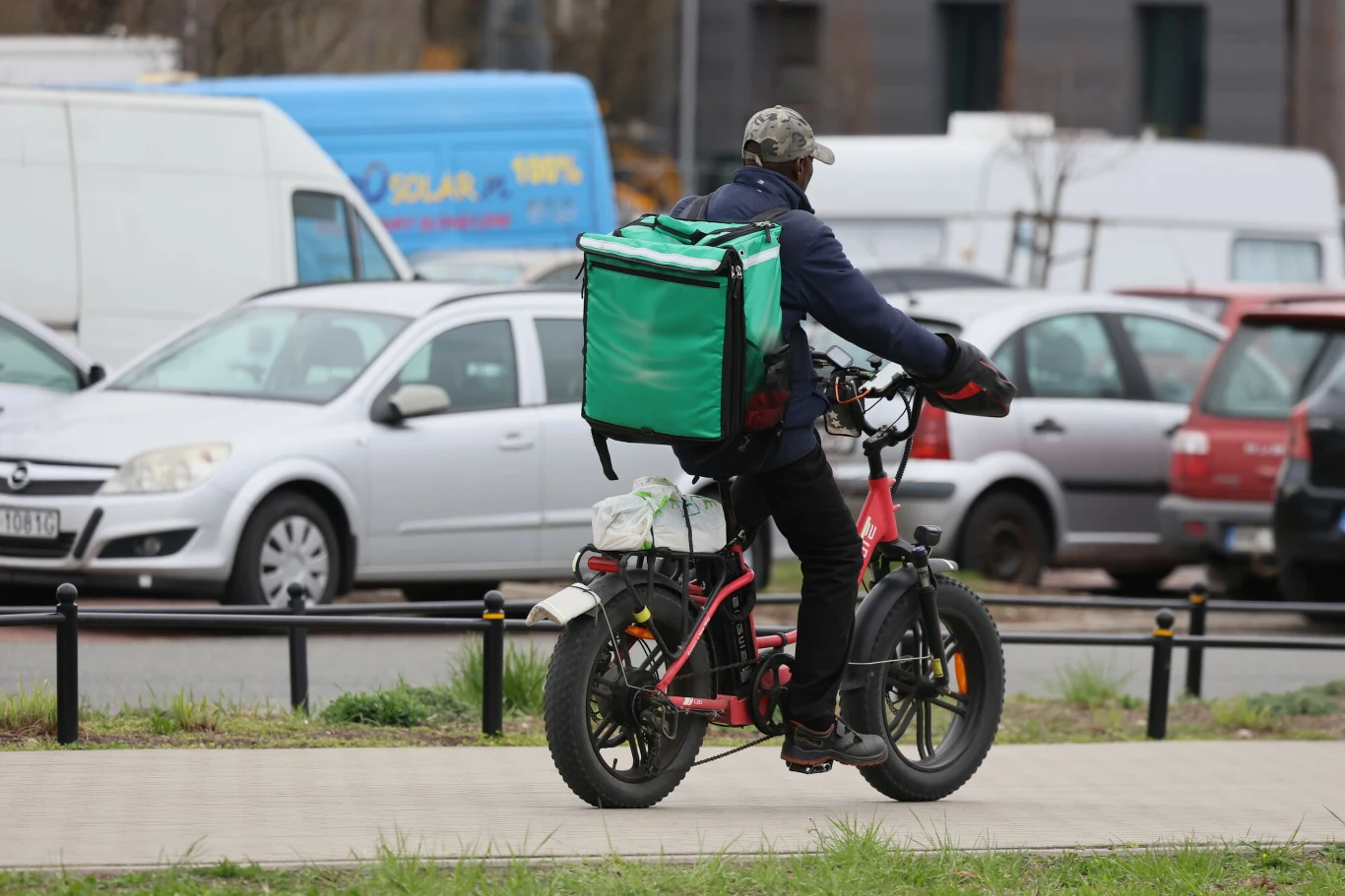 Rowery elektryczne z Chin zalały polskie ulice Dostawca jedzenia jadący elektrycznym rowerem po chodniku, ubrany w niebieską kurtkę i czapkę z daszkiem, z dużą zieloną torbą na plecach. W tle widoczne zaparkowane samochody i furgonetki.