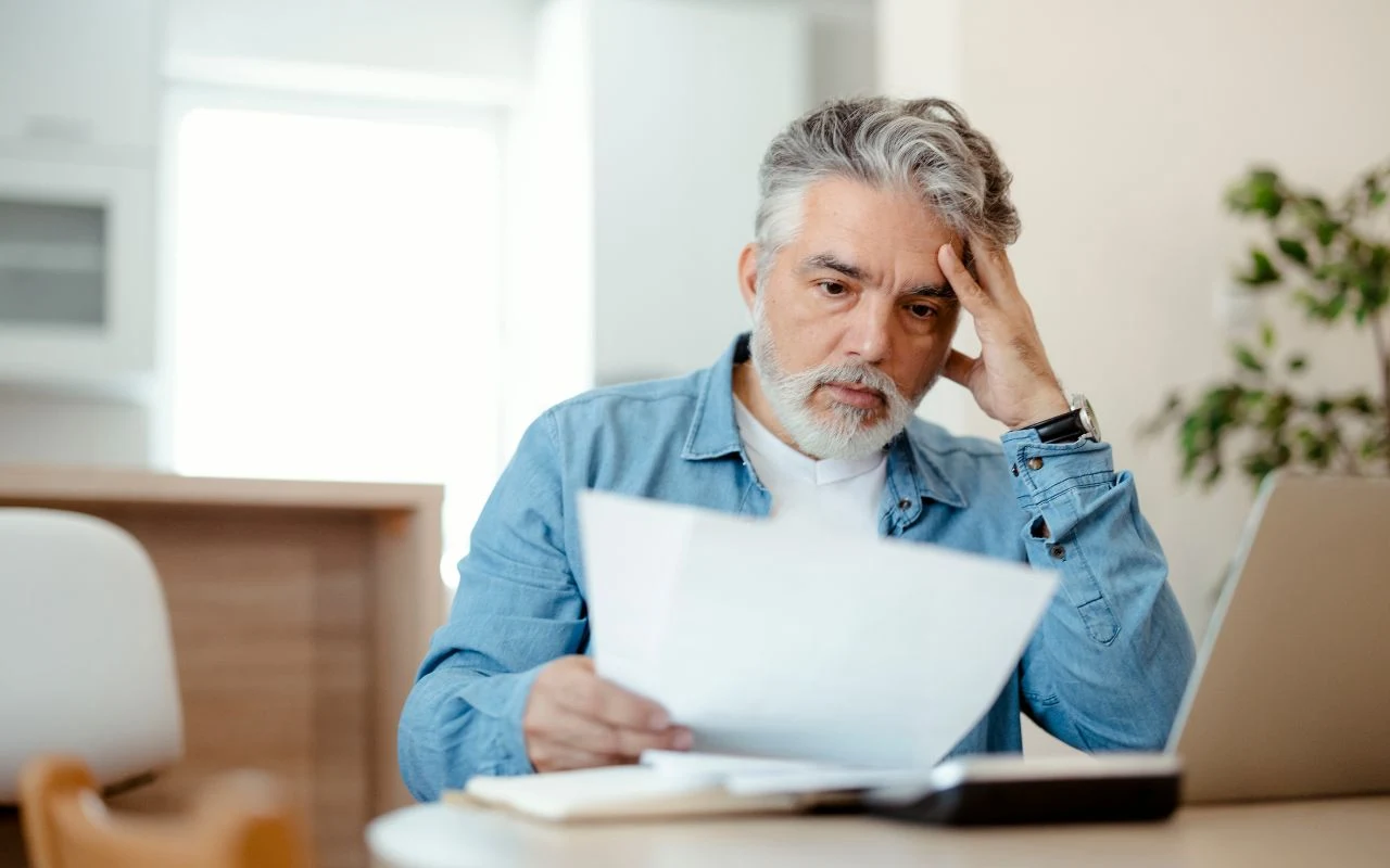 Starszy mężczyzna z siwymi włosami i brodą siedzi przy stole, skupiony na czytaniu dokumentów, opierając głowę na dłoni, przed nim leży otwarty laptop.
