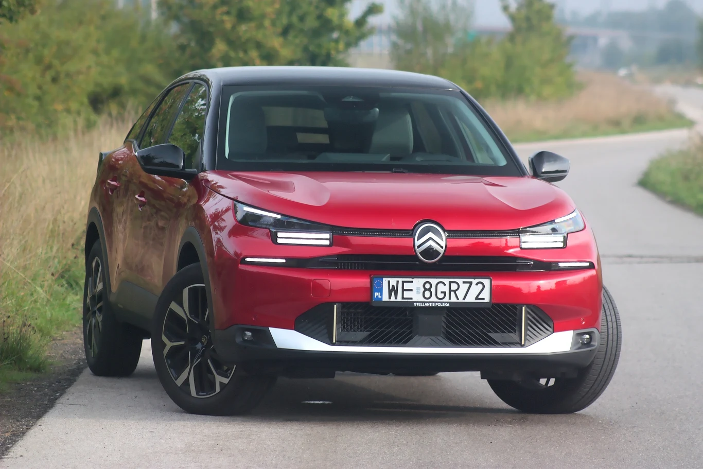Citroen C4 po modernizacji otrzymał mocno zmieniony przód Czerwony samochód Citroën C4 stoi na poboczu wąskiej asfaltowej drogi, otoczonej zielenią i niskimi krzakami, auto widoczne od przodu z wyraźnym numerem rejestracyjnym.