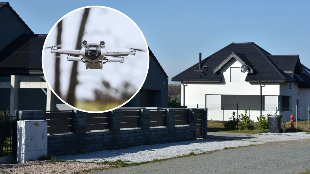 Nowoczesny dom jednorodzinny z jasną elewacją i ciemnym dachem otoczony ogrodzeniem, na pierwszym planie powiększenie lecącego drona typu quadrocopter.
