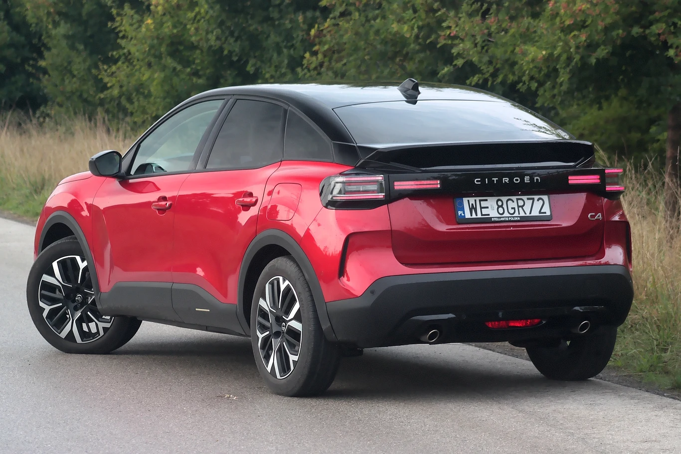 Z tyłu również zmieniono światła, pojawiła się także czarna blenda z napisem PEUGEOT Czerwony samochód Citroën C4 z czarnym dachem zaparkowany przy drodze, widok od tyłu, w tle zieleń i drzewa.