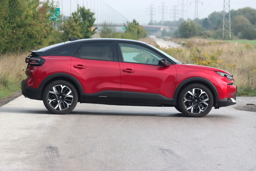 Samochody w najbogatszej wersji Max wyróżniają się ciemnymi szybami tylnymi i srebrną osłoną pod silnikiem Czerwony samochód osobowy nowoczesnego typu zaparkowany poprzecznie na asfaltowej drodze, otoczony zielenią i elementami infrastruktury drogowej.