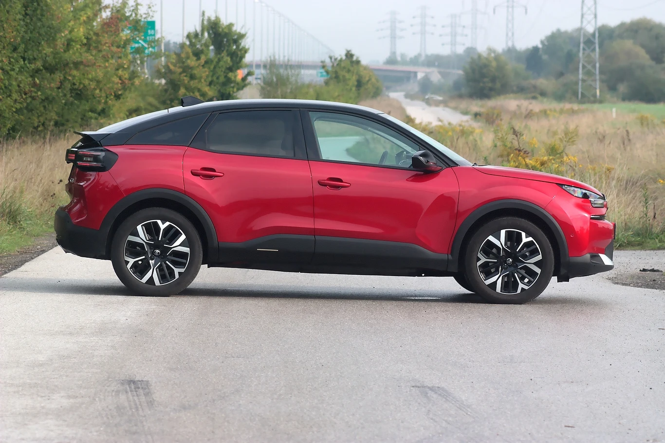 Samochody w najbogatszej wersji Max wyróżniają się ciemnymi szybami tylnymi i srebrną osłoną pod silnikiem Czerwony samochód osobowy nowoczesnego typu zaparkowany poprzecznie na asfaltowej drodze, otoczony zielenią i elementami infrastruktury drogowej.