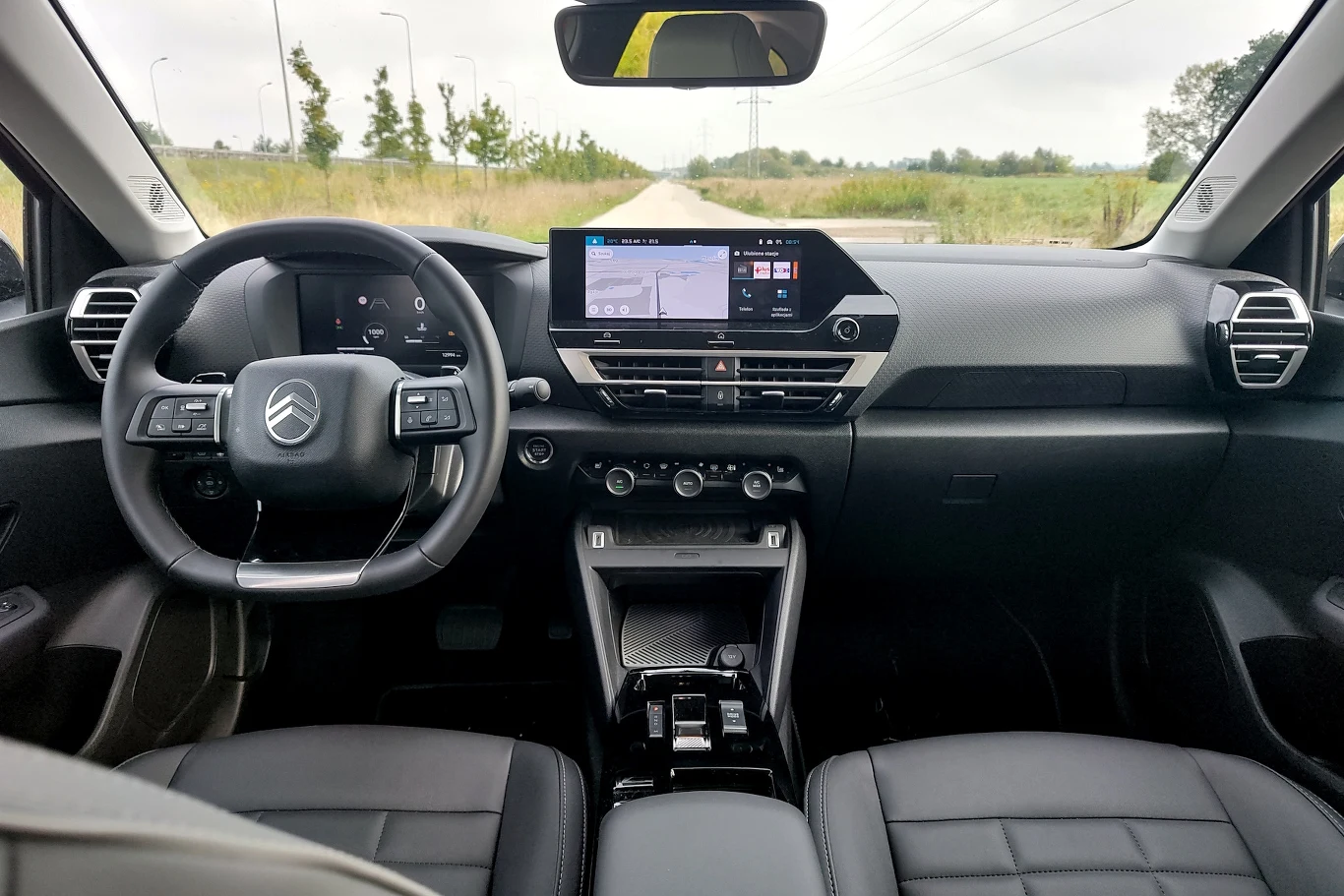 Ekran cyfrowych zegarów nadal ma 10 cali Nowoczesne wnętrze samochodu Citroën z cyfrowym panelem wskaźników, dużym ekranem multimedialnym na konsoli środkowej, wielofunkcyjną kierownicą oraz luksusowymi, czarnymi fotelami skórzanymi. Za szybą widoczna prosta droga prowadząca w stronę zieloneg...