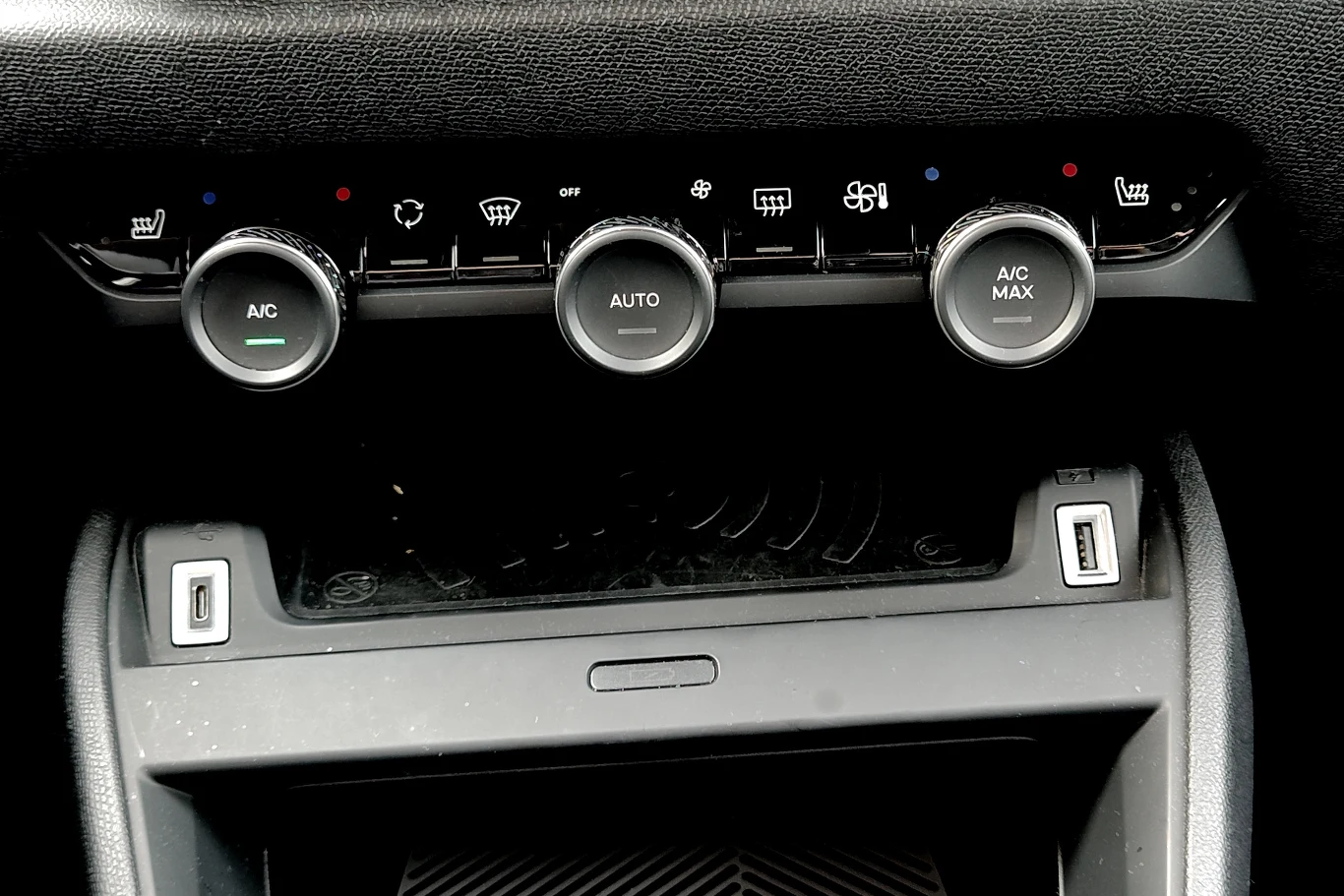Fizyczny panel sterowania klimatyzacją to wygodne rozwiązanie. Pod nim znajduje się ładowarka indukcyjna Panel sterowania klimatyzacją w samochodzie z trzema dużymi pokrętłami oznaczonymi jako A/C, AUTO oraz A/C MAX, poniżej widoczne dwa porty USB.