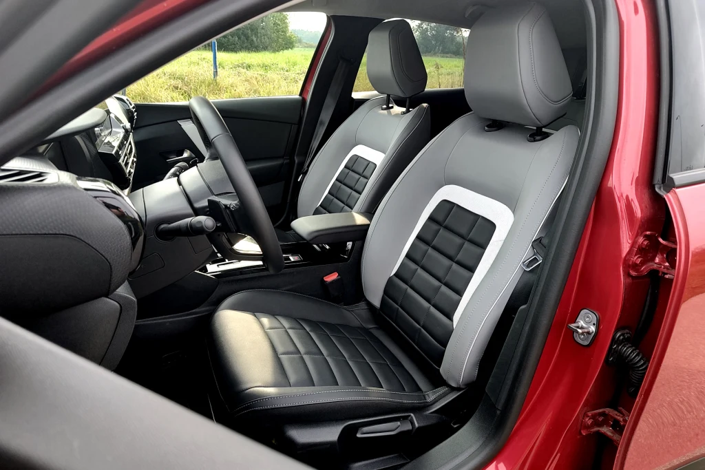 Przy okazji liftingu Citroen C4 otrzymał nowe fotele Wnętrze nowoczesnego samochodu z widocznymi dwoma przednimi siedzeniami, wielofunkcyjną kierownicą i deską rozdzielczą wykończoną w jasnych kolorach, po otwarciu drzwi kierowcy; tapicerka w kontrastowych barwach, elementy czarno-białe.