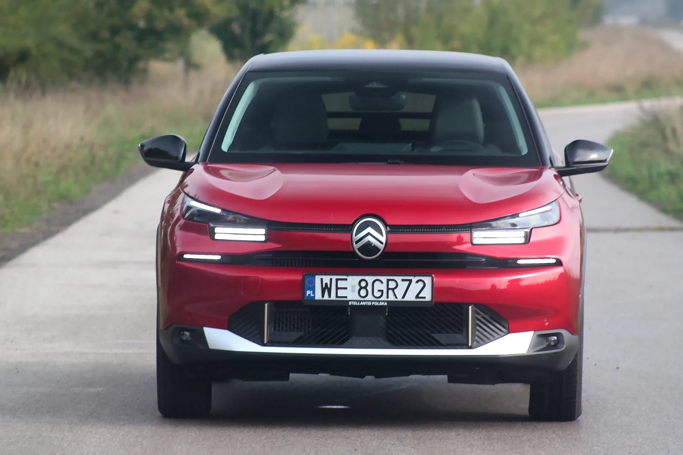 Stylizacja przedniej części C4 po liftingu nie każdemu musi się podobać Nowoczesny czerwony samochód Citroen zaparkowany na bocznej, wiejskiej drodze, widoczny z przodu, z wyraźnie widoczną rejestracją oraz charakterystycznym logo marki.