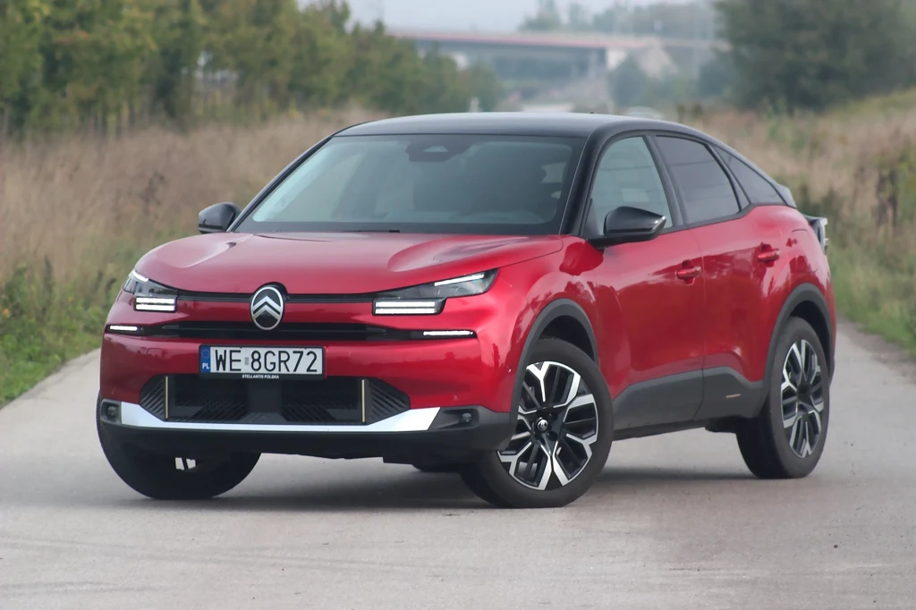 Citroen C4 po liftingu Czerwony samochód Citroën C4 stoi na asfaltowej drodze otoczonej trawą, widoczny z przodu pod lekkim kątem. Samochód posiada nowoczesne światła LED i czarne akcenty nadwozia, a w tle widoczne są drzewa oraz podwyższona droga.