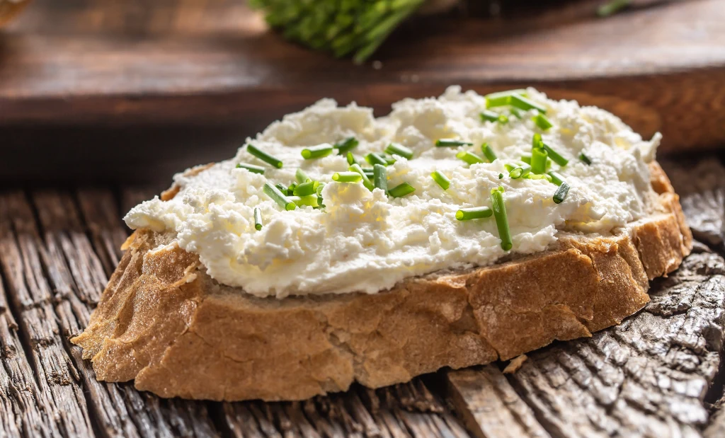 Bryndza ma specyficzny smak, ale istnieją jej miłośnicy Kromka chleba z grubą warstwą białego twarogu, posypana siekanym szczypiorkiem, leży na drewnianym stole.