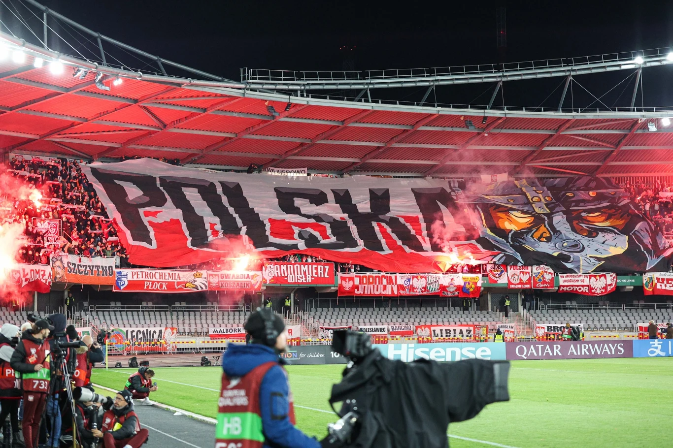 PZPN został ukarany przez FIFA za zachowanie kibiców podczas meczu Litwa - Polska Trybuny stadionu wypełnione kibicami, ogromna flaga z napisem 'Polska' i artystycznym motywem wilka, czerwień rac dymnych, a na pierwszym planie osoby z kamerami dokumentujące wydarzenie.