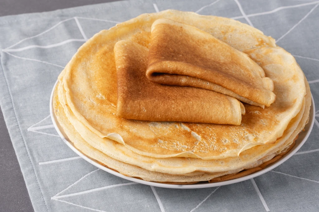 Stos cienkich, złocistych naleśników ułożonych na białym talerzu, na wierzchu dwa naleśniki są złożone w trójkąty. Całość ustawiona na szarej serwetce z białym geometrycznym wzorem.