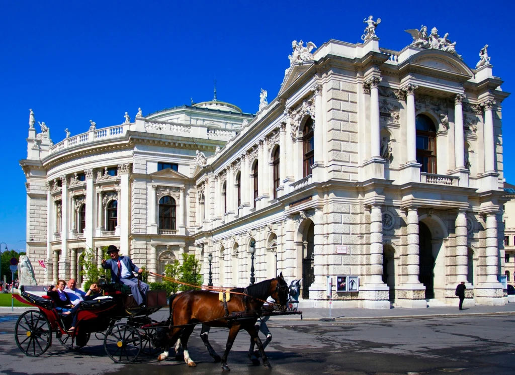 Imponujący budynek w stylu klasycystycznym ozdobiony licznymi kolumnami, rzeźbami i łukami, przed którym przejeżdża bryczka zaprzężona w dwa konie z jadącymi pasażerami oraz powożącym woźnicą ubranym w strój galowy.