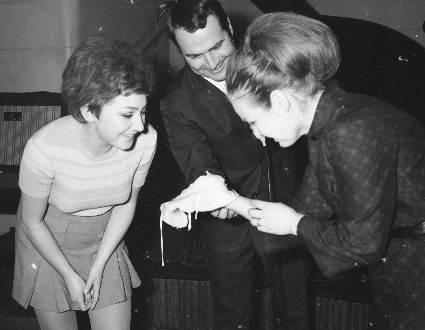 Troje ludzi bierze udział w tradycyjnej zabawie andrzejkowej polegającej na laniu wosku przez klucz. Kobieta w sukience trzyma świeczkę nad naczyniem z wodą, podczas gdy pozostałe osoby uważnie obserwują wynik lania wosku.