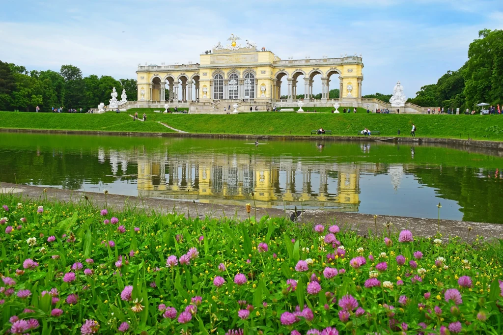 Okazały budynek Glorietty odbijający się w wodzie stawu, w otoczeniu bujnej zieleni z kolorowymi kwiatami koniczyny na pierwszym planie, a w tle widać spacerujących ludzi i rzeźby ustawione po bokach alei.
