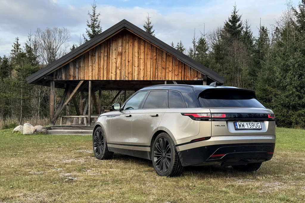 Luksusowy SUV w kolorze srebrnym zaparkowany na trawie przed drewnianą altaną otoczoną lasem iglastym, widoczne tablice rejestracyjne, spokojna wiejska sceneria.