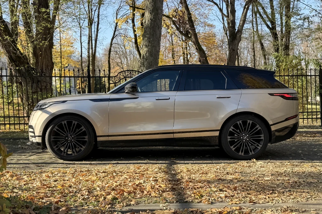 Luksusowy SUV w kolorze srebrnym stoi zaparkowany na tle jesiennych drzew i czarnego metalowego ogrodzenia, pojazd na dużych, czarnych felgach, otoczenie pokryte suchymi liśćmi.