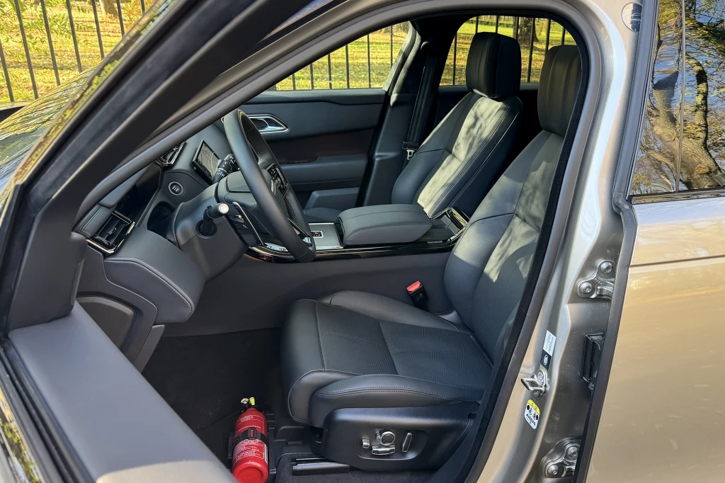 Wnętrze nowoczesnego samochodu z widocznymi czarnymi skórzanymi siedzeniami, kierownicą, deską rozdzielczą i zamontowaną przy podłodze gaśnicą po stronie pasażera.