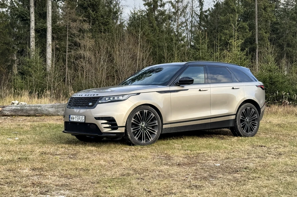 Luksusowy SUV Range Rover Velar w beżowym kolorze stoi zaparkowany na trawie, otoczony przez las iglasty. Auto charakteryzuje się nowoczesnym designem, dużymi felgami i przyciemnianymi szybami.