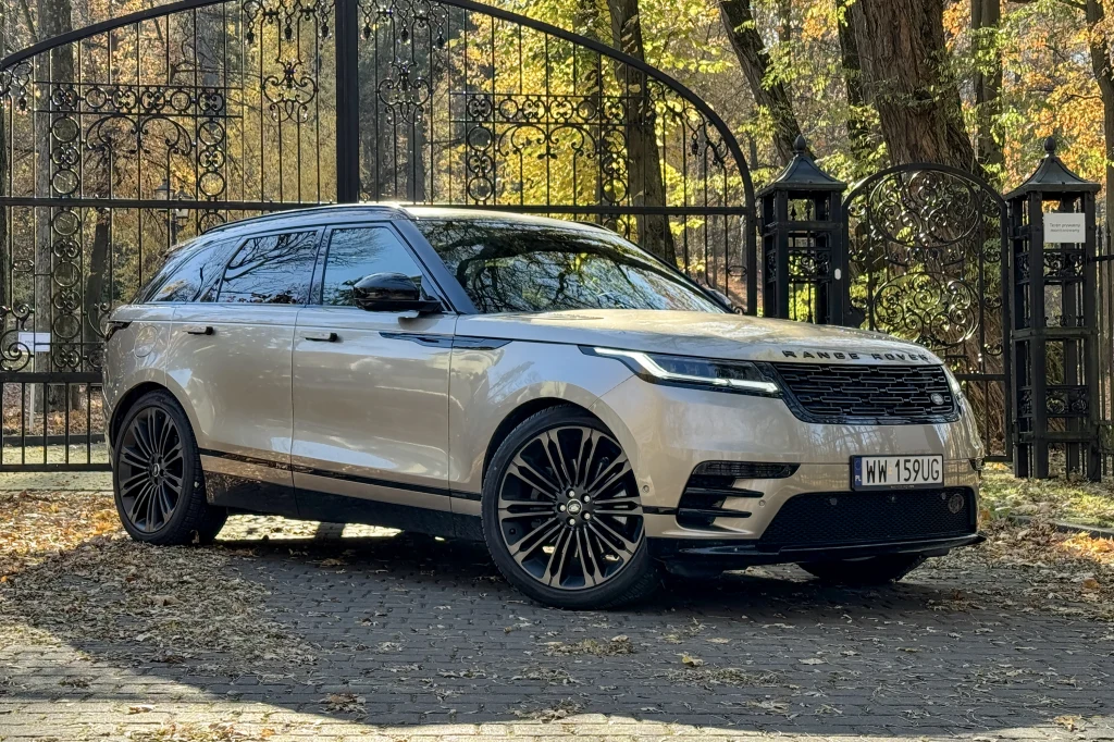 Luksusowy samochód terenowy Range Rover Velar zaparkowany na tle ozdobnej bramy z kutego żelaza w otoczeniu jesiennych drzew.