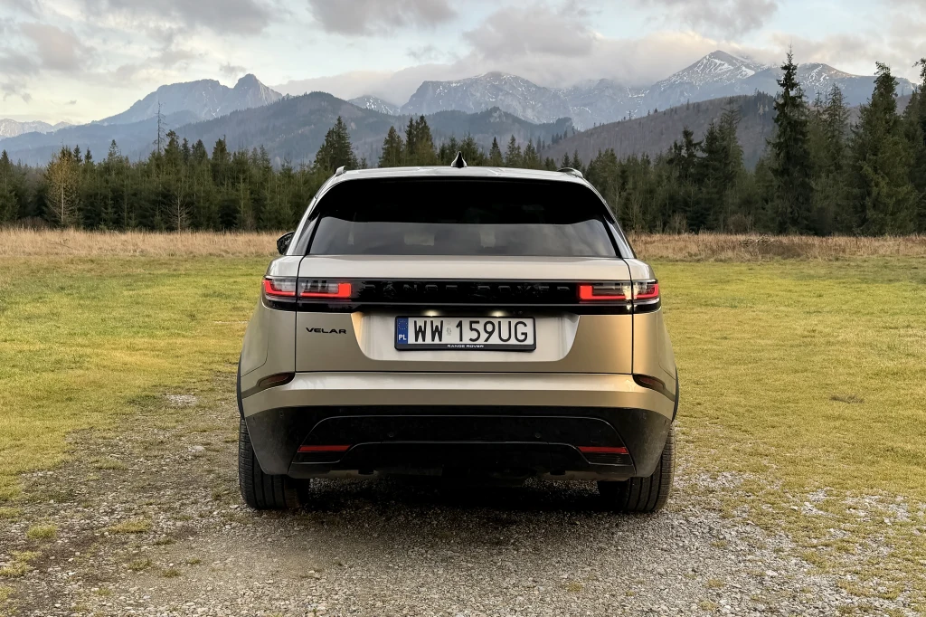 Samochód terenowy marki Range Rover Velar zaparkowany tyłem na zielonej łące z widokiem na górski krajobraz, otoczony sosnami i w oddali ośnieżonymi szczytami.