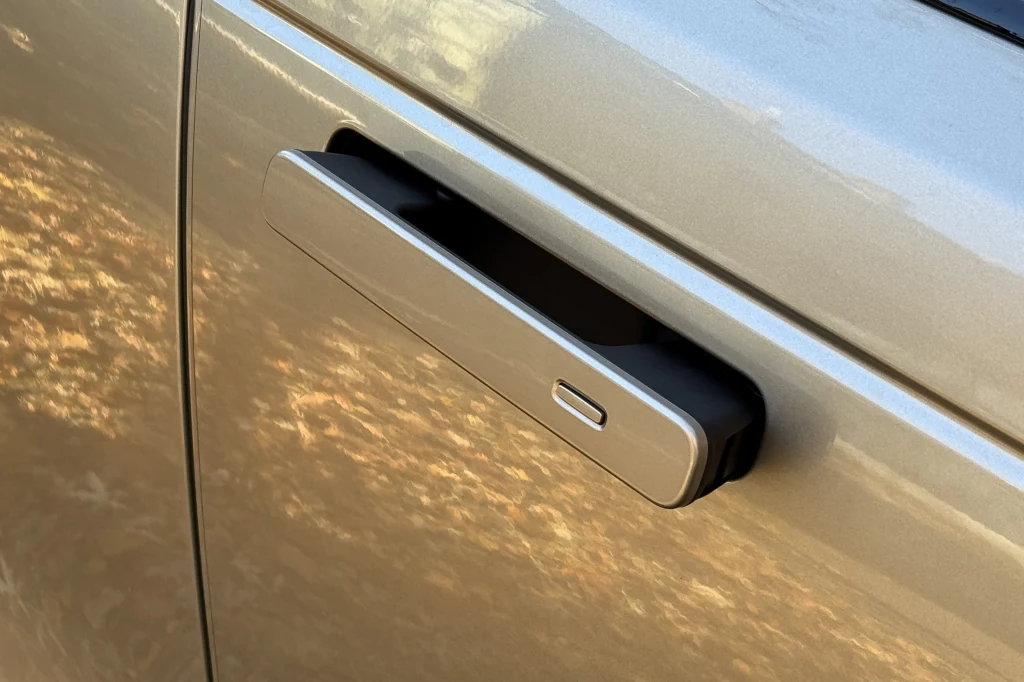 Drzwi samochodu z wysuniętą nowoczesną klamką w kolorze srebrnym, na tle jasnej metalicznej karoserii.