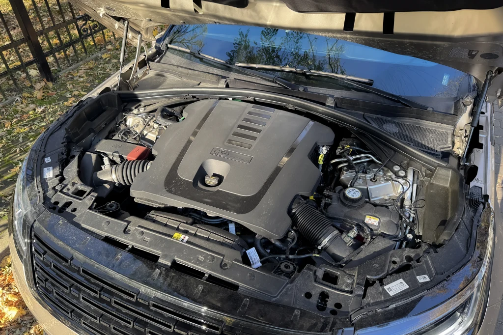 Silnik samochodowy widoczny pod otwartą maską, w otoczeniu części mechanicznych oraz przewodów, z elementami wykonanymi głównie z metalu i tworzyw sztucznych. Samochód stoi zaparkowany na zewnątrz, widać opadłe liście na ziemi oraz metalowe ogrodzenie ...