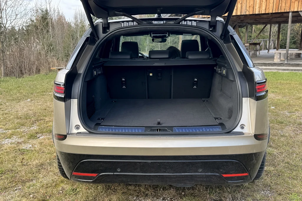 Otwarty bagażnik dużego samochodu typu SUV, wnętrze przestronne, czyste, tylne siedzenia widoczne poprzez duży otwór załadunkowy, auto zaparkowane na trawie przy drewnianej altanie.