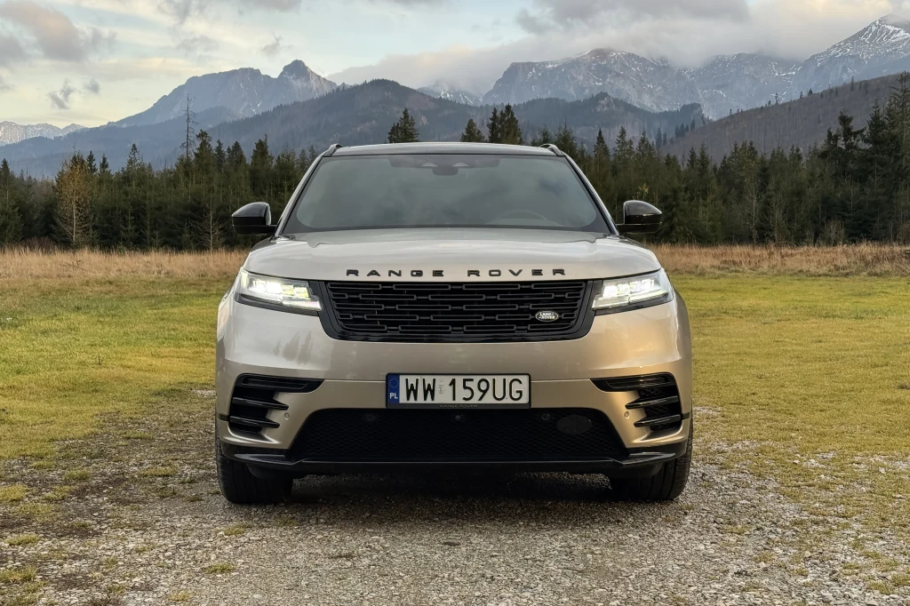 Luksusowy samochód Range Rover stoi zaparkowany na tle gór i lasu. Widok z przodu ukazuje nowoczesny design pojazdu oraz elegancki grill. W tle rozpościera się panorama gór ze szczytami częściowo zakrytymi chmurami.