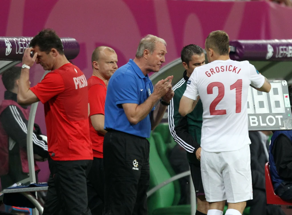 Zmiana zawodnika podczas meczu piłkarskiego, trener rozmawia z piłkarzem w białej koszulce z numerem 21, obok stoją inni członkowie sztabu oraz piłkarz w czerwonej koszulce.