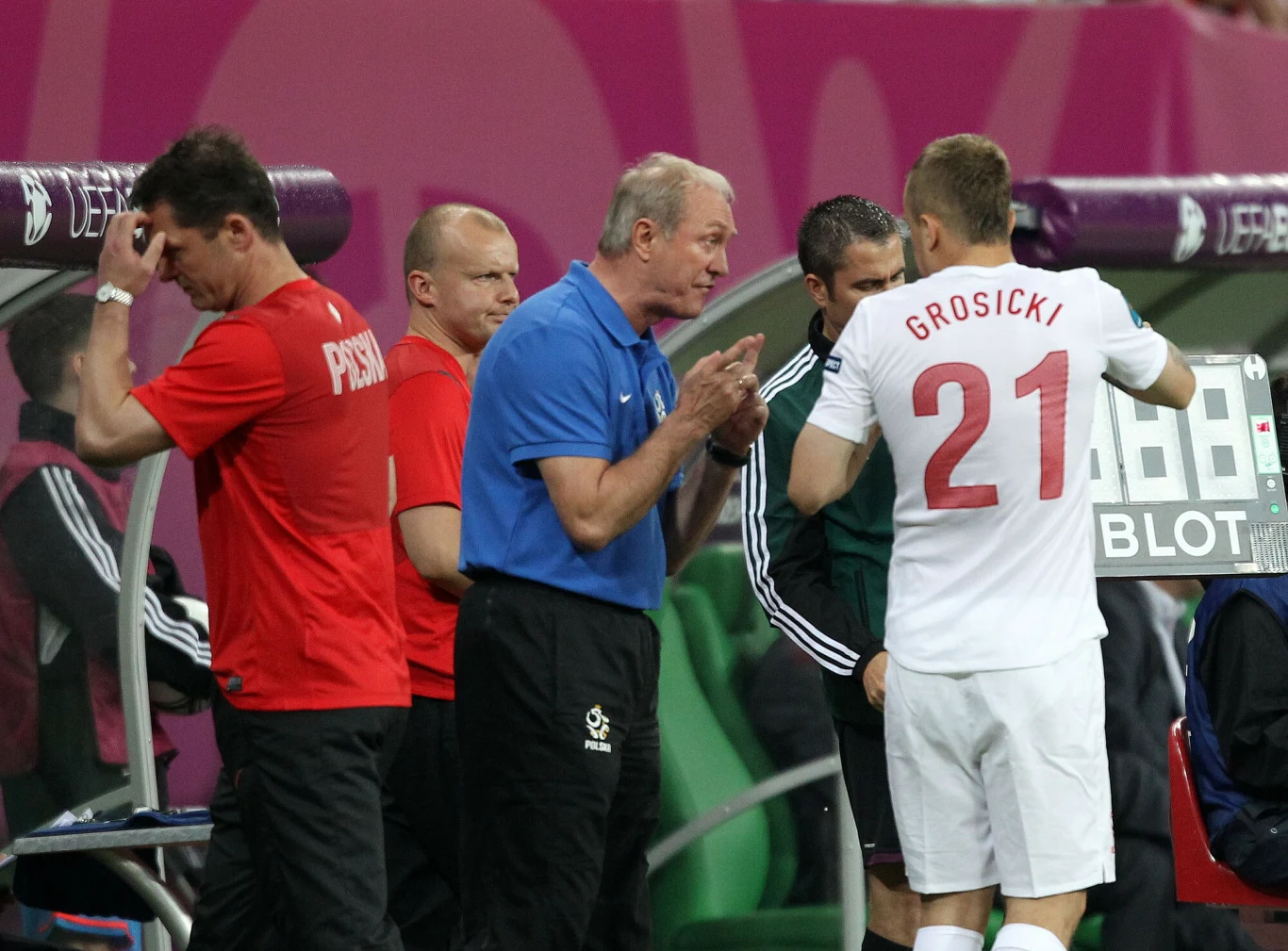 Zmiana zawodnika podczas meczu piłkarskiego, trener rozmawia z piłkarzem w białej koszulce z numerem 21, obok stoją inni członkowie sztabu oraz piłkarz w czerwonej koszulce.