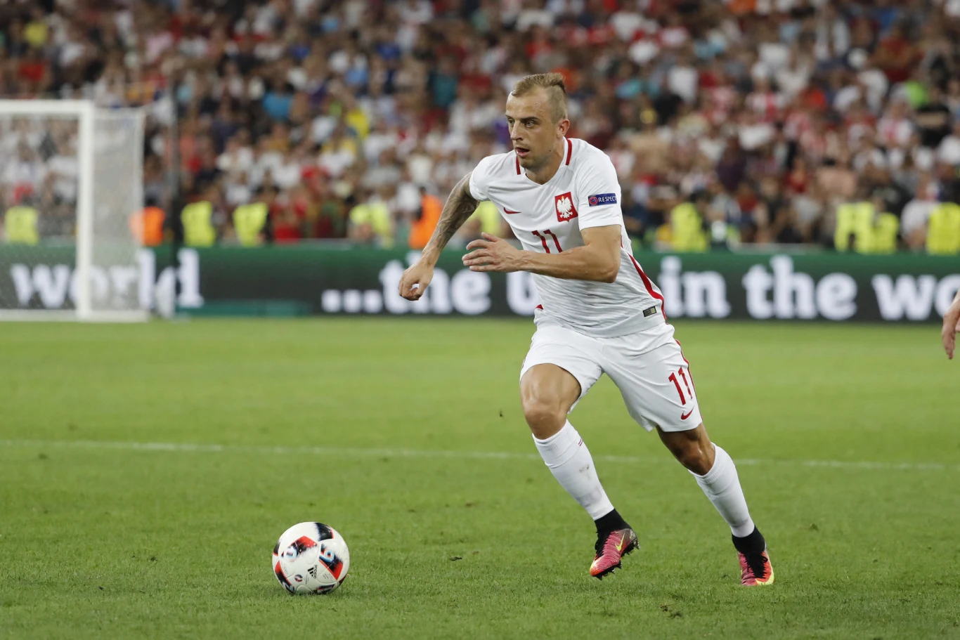 Piłkarz reprezentacji Polski w białym stroju z numerem 11 biegnie za piłką podczas meczu piłkarskiego na stadionie, w tle trybuny wypełnione kibicami.