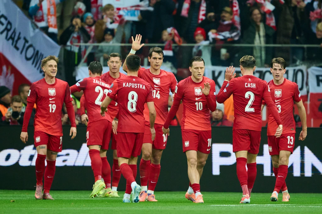 Polscy piłkarze w czerwonych strojach świętują zdobycie bramki lub ważny moment podczas meczu na stadionie, otoczeni kibicami z polskimi flagami.