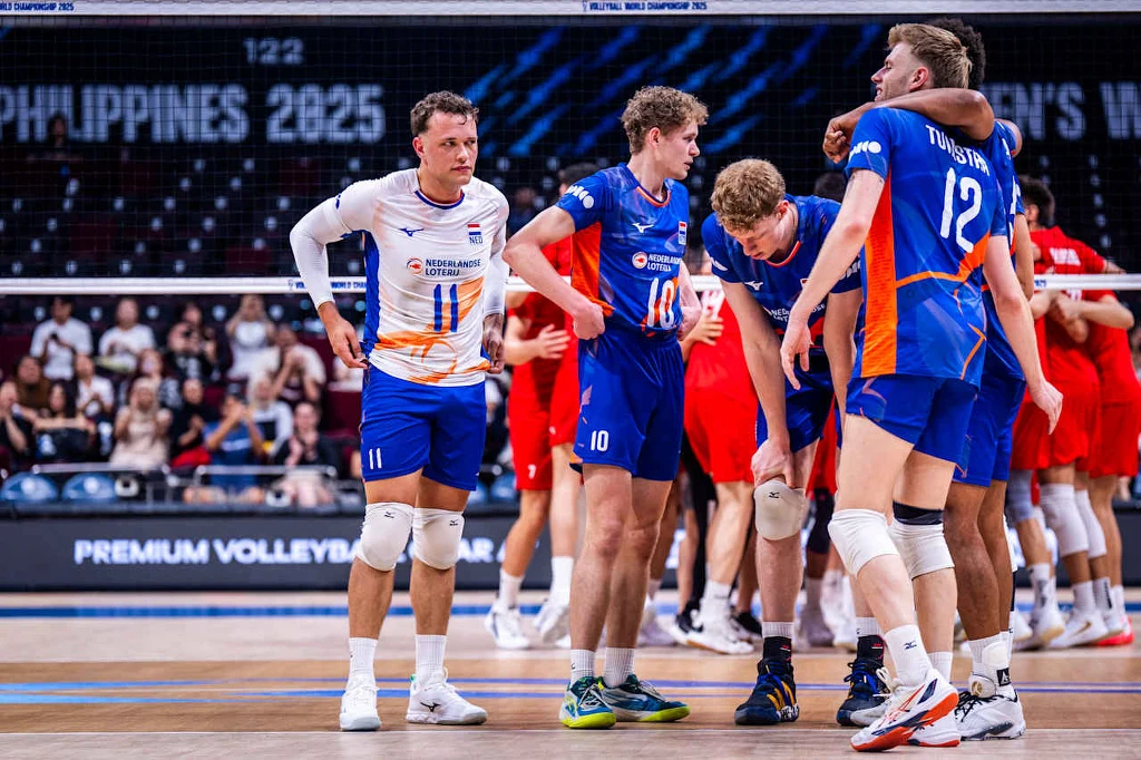 Siatkarze reprezentacji Holandii, z nr 12 Bennie Tuinstra Zawodnicy męskiej drużyny siatkarskiej w niebiesko-pomarańczowych strojach omawiają taktykę na boisku, podczas meczu przeciwko drużynie w czerwonych strojach, przy widocznych trybunach w tle.