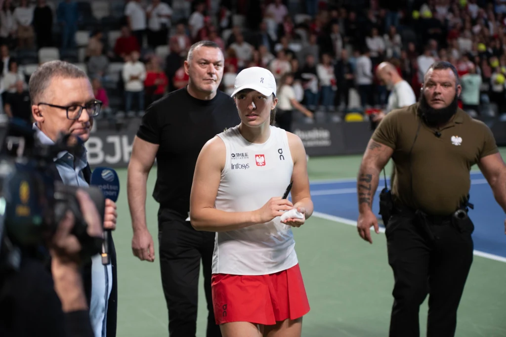 Tenisistka w białej czapce i białym topie z czerwonymi spodenkami stoi na korcie tenisowym, otoczona przez ochroniarzy i dziennikarzy. W tle widoczna publiczność na trybunach.