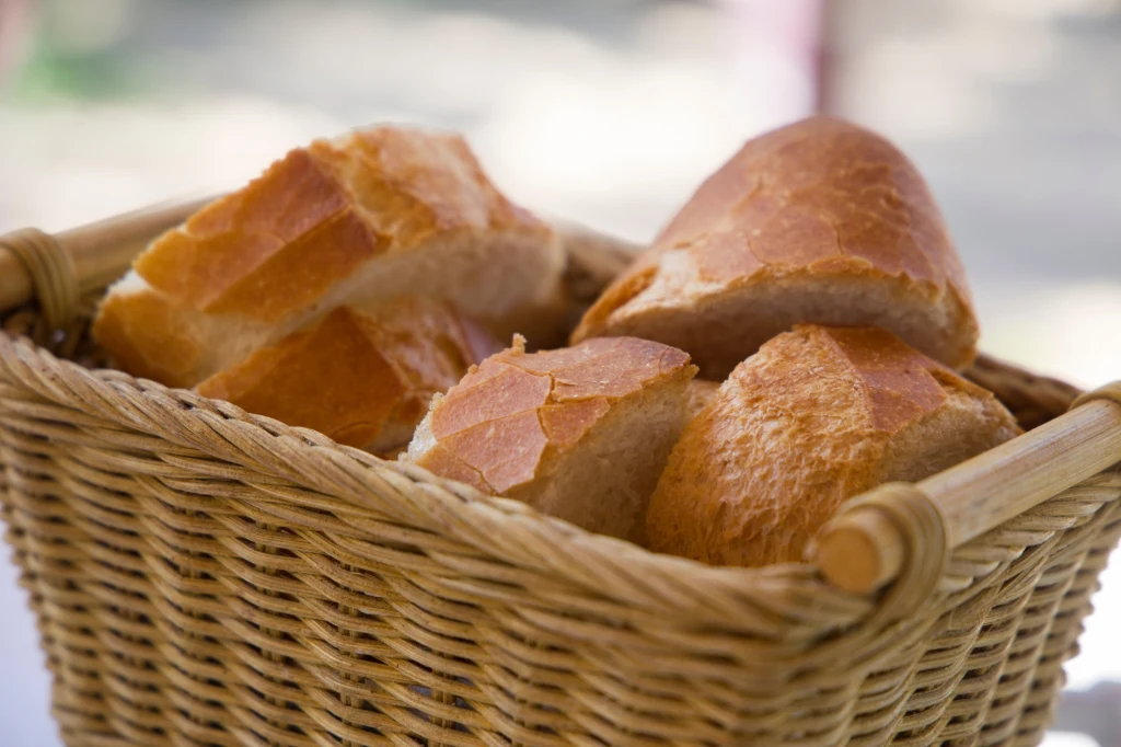 Co zrobić z czerstwego chleba? Kosz z wikliny wypełniony świeżymi, chrupiącymi bułkami z jasną i lekko błyszczącą skórką, ułożonymi na białym tle.