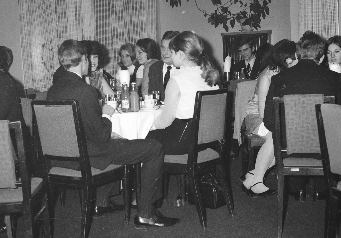 Grupa młodych osób w eleganckich ubraniach siedzi przy kilku stolikach w restauracji lub kawiarni, prowadząc rozmowy i spędzając wspólnie czas. Na stołach stoją butelki z napojami i szklanki, a w tle widać zasłony oraz roślinność.