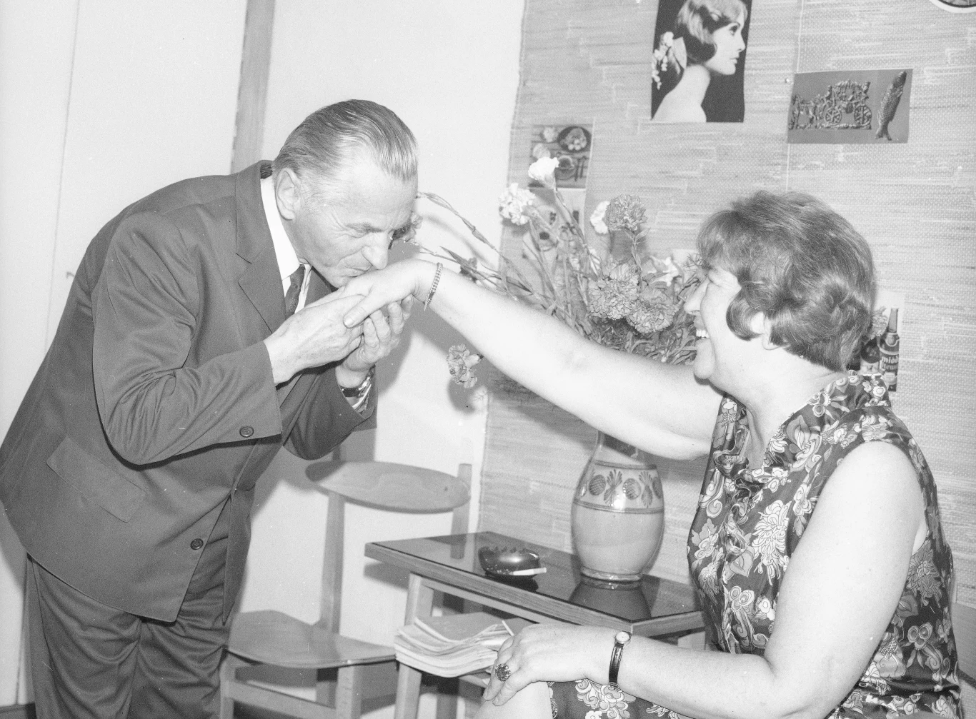 Starszy mężczyzna w garniturze całuje dłoń uśmiechniętej kobiety siedzącej na krześle, w tle wazon z kwiatami na stoliku oraz zdjęcia i grafiki zawieszone na ścianie.