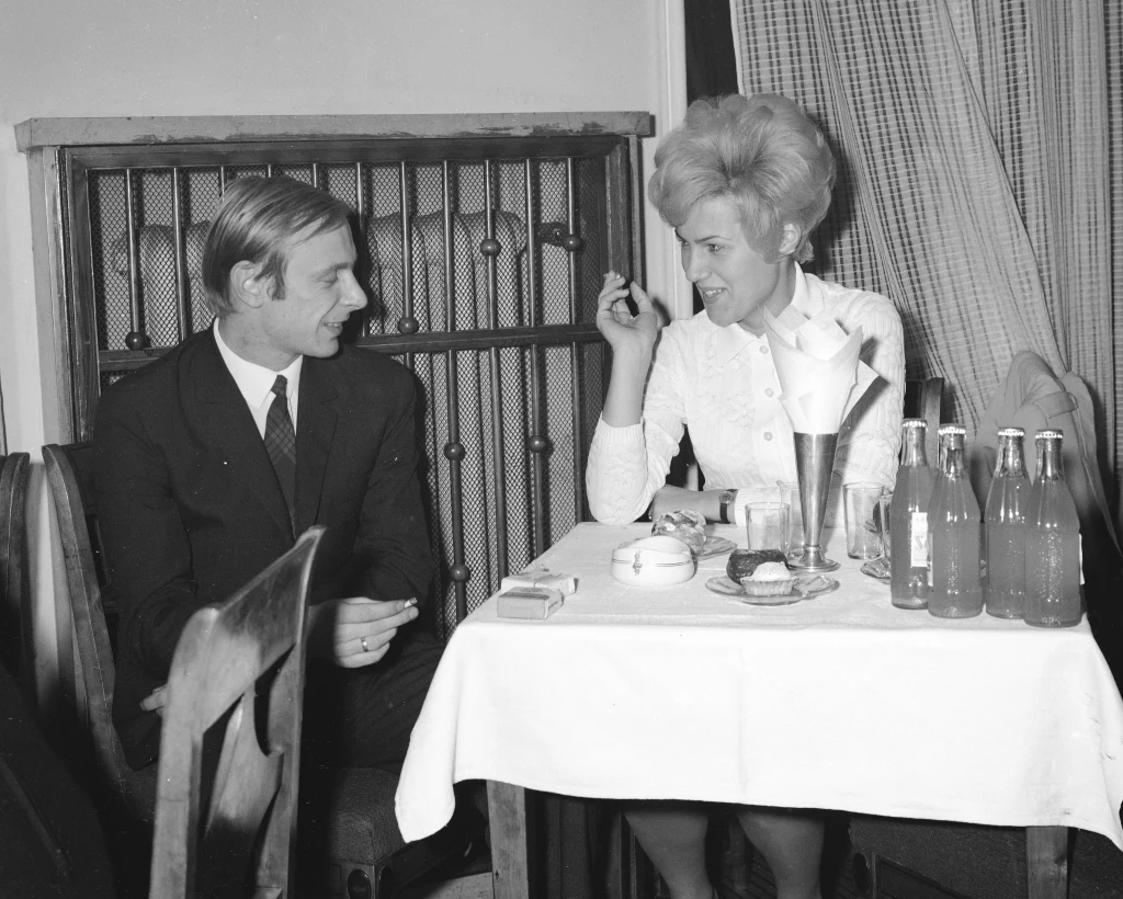 Andrzejki w kawiarni "Nowy Świat", lata 60. Kobieta i mężczyzna w eleganckich ubraniach siedzą przy stole nakrytym białym obrusem, na którym stoją butelki napojów i filiżanki. Oboje rozmawiają i uśmiechają się, tworząc przyjazną atmosferę spotkania towarzyskiego.