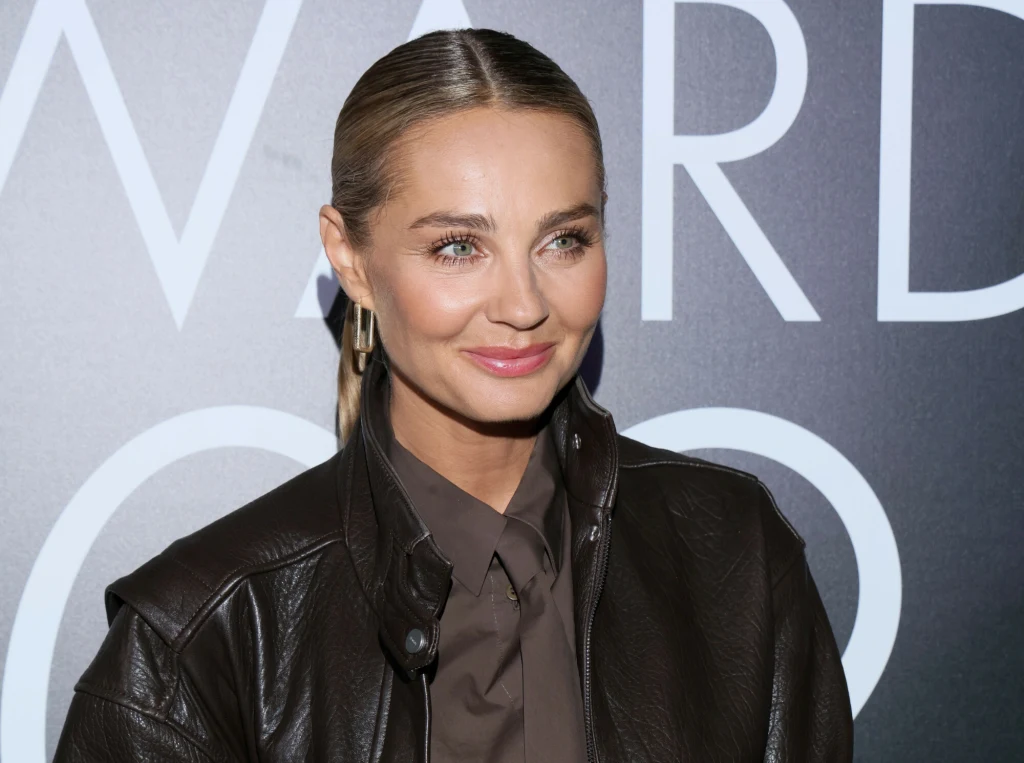 Kobieta o blond włosach związanych w gładki kucyk, ubrana w ciemną koszulę i czarną skórzaną kurtkę, stoi na tle grafiki z dużymi białymi literami.