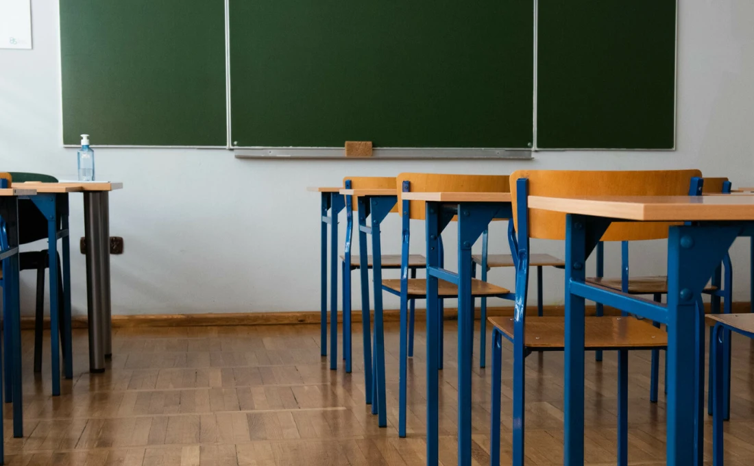 Gójsk. Atak rodzica na dyrektorkę szkoły (Zdjęcie ilustracyjne) Puste, drewniane ławki ustawione w rzędach na podłodze z paneli, w tle duża zielona tablica oraz butelka płynu dezynfekującego na metalowym stoliku po lewej stronie.